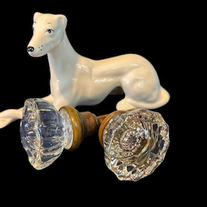 May include: Two antique glass door knobs with a decorative, faceted design. The knobs are attached to wooden spindles. A white porcelain figurine of a dog is lying on top of the knobs.