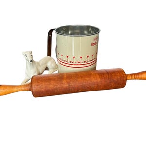 Può includere: Un mattarello di legno con un manico marrone e una statuetta di ceramica bianca di un cane sullo sfondo. È anche visibile un misurino in smalto bianco con un motivo floreale rosso e bianco e il testo "Dipinto a mano".