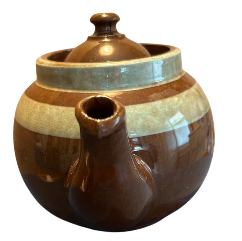 May include: A brown ceramic teapot with a lid and spout. The teapot has horizontal stripes in shades of brown and cream, with a light blue band around the top. The handle is not visible in the image.