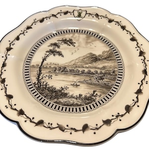 Può includere: Un piatto decorativo bianco sporco con bordo smerlato e bordo nero. Il piatto presenta una scena paesaggistica color seppia al centro, circondata da un bordo dettagliato. Un motivo di foglie e viti marrone scuro adorna il bordo. Un piccolo stemma in alto.