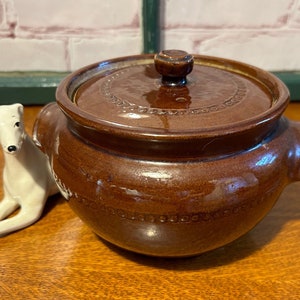 May include: A brown ceramic pot with a lid and two handles. The pot has a decorative design around the rim.