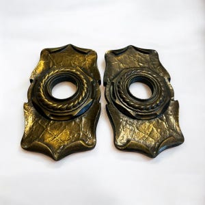 May include: Two antique bronze-colored cabinet pulls. Each pull has a decorative, textured backplate with a central circular element featuring a rope-like design around a central hole. The pulls have a vintage aesthetic.