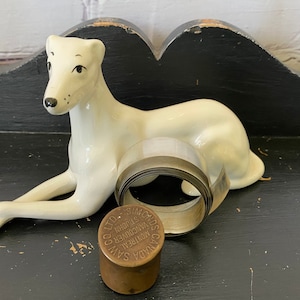 Può includere: Una statuetta di ceramica bianca di un cane sdraiato con un rotolo di nastro adesivo trasparente e una bobina di ottone con il testo "CANADA SAW & TOOL CO. LTD. MONTREAL VANCOUVER ST. JOHN" su di essa.