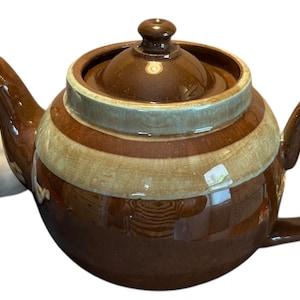May include: A brown ceramic teapot with a lid and handle, featuring horizontal stripes in shades of brown and beige. A white ceramic dog figurine is positioned to the left of the teapot.