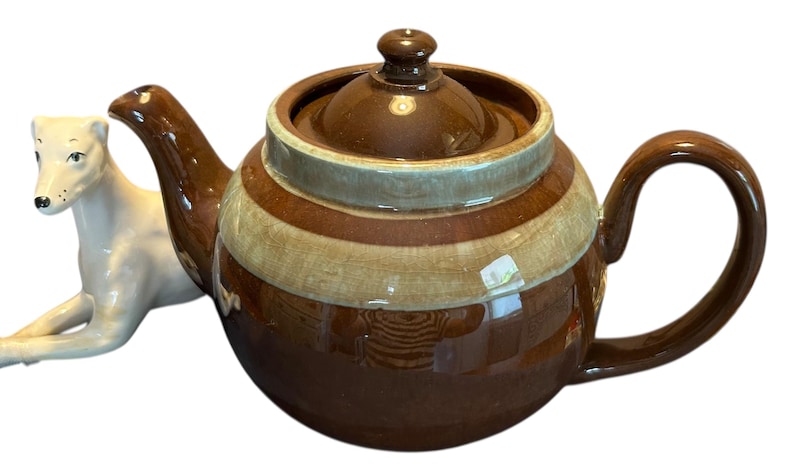 May include: A brown ceramic teapot with a lid and handle, featuring horizontal stripes in shades of brown and beige. A white ceramic dog figurine is positioned to the left of the teapot.