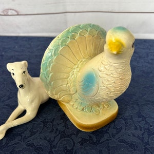 Peut inclure: Figurine de dinde en céramique avec un corps crème, une queue bleue et verte et un bec jaune. Une figurine de chien crème est allongée à côté de la dinde. Les figurines sont sur une surface bleu foncé.