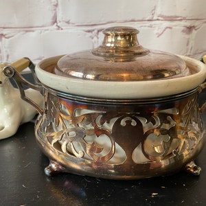 K&ouml;nnte beinhalten: Eine silberne und wei&szlig;e Keramik-Servierplatte mit Deckel und verziertem Design. Die Platte hat einen dekorativen, durchbrochenen Sockel und zwei Griffe.