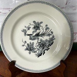 Op de afbeelding: Een witte keramische plaat met een grijs en zwart vogel- en bloemmotief. De plaat heeft een grijs patroonrand. De vogel zit tussen bloemen en takken. De plaat wordt op een houten standaard gepresenteerd.