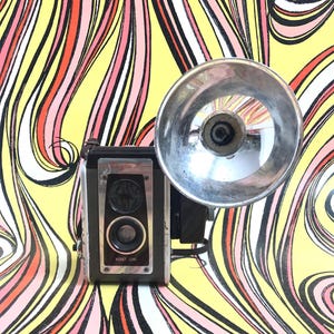 May include: Vintage black and silver Kodak Brownie camera with a flash attachment. The camera has a silver lens and the words "KODET LENS" are printed on the front. The camera is set against a colorful, abstract background.