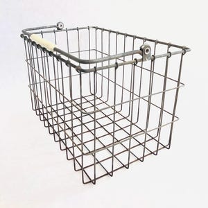 May include: A rectangular wire basket with a metal frame and a light-colored wooden handle. The basket is made of dark gray metal and has a grid-like pattern. The handle is attached to the top of the basket with metal brackets.