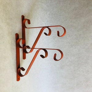 May include: Two rust-colored metal plant hangers with decorative swirls. The hangers are attached to a wall.