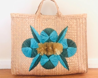 May include: Woven tote bag with a rectangular shape and two handles. The bag is beige with a decorative floral design in teal, dark green, and gold. The bag is sitting on a wooden surface.