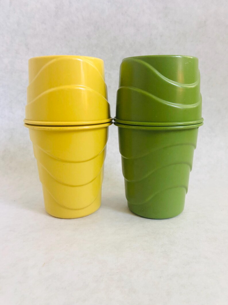 Puede incluir: Dos vasos apilables vintage, uno amarillo brillante y otro verde. Ambos tienen un dise&ntilde;o ondulado y acanalado, con una banda met&aacute;lica en el medio. Los vasos probablemente son de pl&aacute;stico.