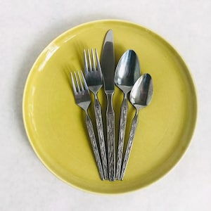 May include: A set of five silver-toned utensils with textured handles, including a knife, fork, and three spoons, arranged on a yellow plate.