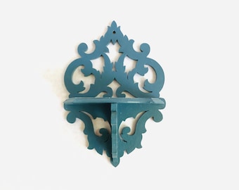 May include: A teal blue wall shelf with a decorative, ornate design. The shelf is made of wood and has a single shelf.