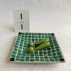 May include: A square, turquoise mosaic tile dish with a border. Two green, elongated objects and two round green objects rest on the dish. A white card with the number "1" is propped up behind the dish. The card reads "MADE IN U.S.A."