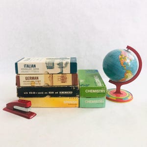 May include: A collection of vintage educational items. Includes language vocabulary card boxes labeled Italian, German, and Spanish, along with chemistry boxes and a small globe. A red stapler is also present.
