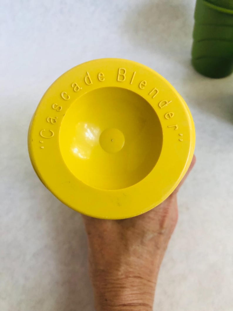 Puede incluir: Tapa de licuadora de color amarillo brillante, con las palabras "Cascade Blender" grabadas en el borde. La tapa es circular con un centro empotrado y est&aacute; sujeta por una mano.