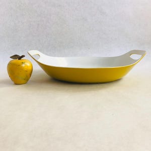 May include: A yellow serving dish with white interior and handle cutouts, next to a yellow apple figurine with a bronze leaf. The dish is a bright yellow color, and the apple figurine has a bronze leaf on top.
