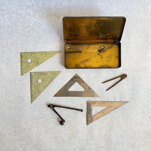 May include: A collection of vintage drafting tools including three triangular rulers, two metal compasses, and a rectangular metal storage tin. The tools are made of brass and steel, featuring etched measurement markings and circular cutouts.
