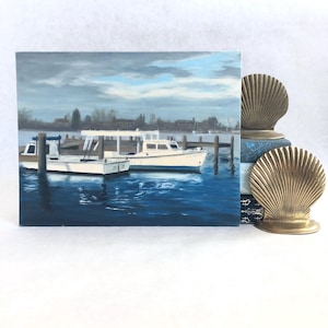 May include: An oil painting depicts two white boats docked in a harbor with blue water under a cloudy sky. The painting is propped up by a stack of books and two gold-colored seashell bookends.