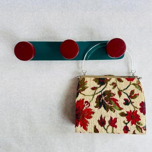 May include: A small, vintage handbag with a floral tapestry design in red, green, and cream hangs from a teal wall hook with three red knobs. The bag has clear plastic handles and a silver clasp.