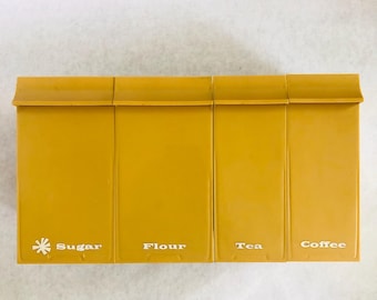 May include: Mustard yellow kitchen canisters labeled Sugar, Flour, Tea, and Coffee. The canisters have a rectangular shape with a slightly curved top and white lettering. A white floral design is next to the word Sugar.