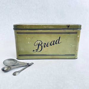 May include: A rectangular olive green metal bread box with a black script label reading Bread and thin black horizontal border lines, accompanied by a set of three small, weathered silver-toned metal measuring spoons attached to a metal ring.