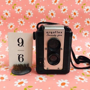 May include: A vintage Argoflex Seventy-five camera with a black body and silver faceplate. The camera is displayed with a white card showing the numbers 9 and 6, and a small floral pin holder. The background is a pink floral pattern.
