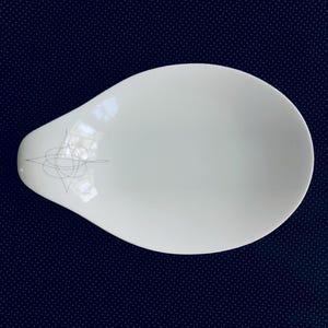 May include: A white, teardrop-shaped serving dish with a subtle, gray geometric design near the handle. The dish has a smooth, glossy surface and is set against a dark blue background.