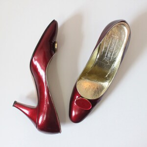 Puede incluir: Un par de zapatos de tacón burdeos brillantes con tacón bajo. Los zapatos tienen punta afilada y un pequeño adorno ovalado en la pala. El interior es dorado.