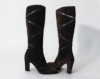 May include: A pair of dark brown suede knee-high boots. The boots have a block heel and a decorative crisscross design on the front. The boots are set against a white background.