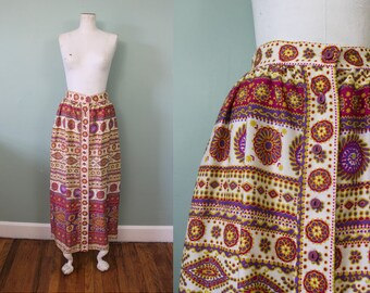 May include: A vintage button-down skirt with a vibrant floral and geometric pattern in shades of white, purple, yellow, and brown. The skirt is displayed on a mannequin and features a high waist and a long, straight silhouette, perfect for a retro look.