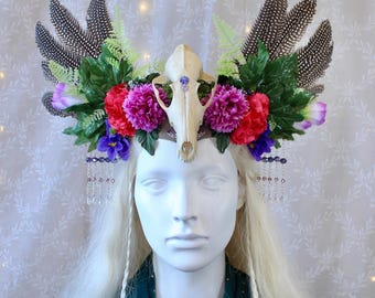 May include: A white skull with purple and pink flowers, green leaves, and feathers in a crown. The crown is on a white mannequin head with long blonde hair.