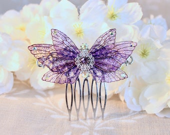 May include: Purple glitter butterfly hair comb with a silver filigree design and a purple gem in the centre. The comb is set against a background of white flowers.