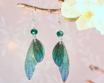May include: Dangle earrings with blue-green iridescent fairy wing charms. Each earring has a green bead and a silver hook. The wings have a glittery finish, and are suspended from a silver wire.