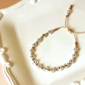 Puede incluir: Un brazalete de ojo turco blanco y dorado con cierre de cord&oacute;n marr&oacute;n. El brazalete est&aacute; hecho de peque&ntilde;as cuentas redondas con un dise&ntilde;o de ojo turco blanco y dorado.