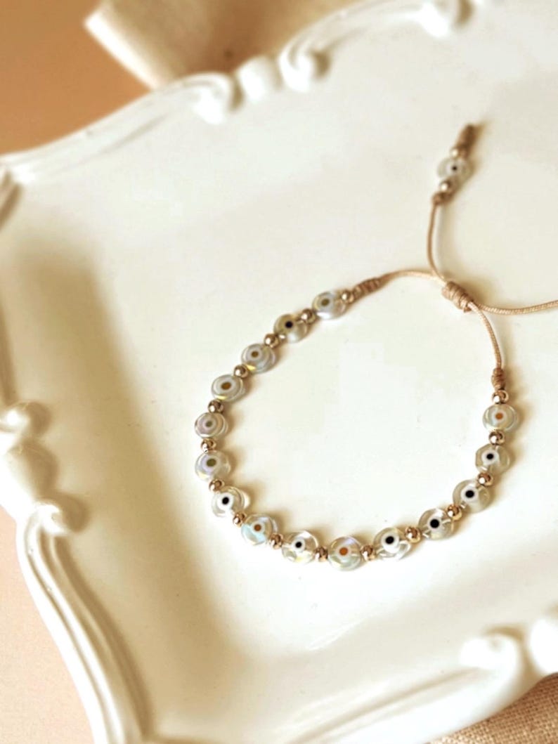Puede incluir: Un brazalete de ojo turco blanco y dorado con cierre de cord&oacute;n marr&oacute;n. El brazalete est&aacute; hecho de peque&ntilde;as cuentas redondas con un dise&ntilde;o de ojo turco blanco y dorado.