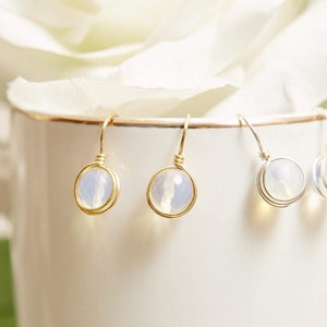 May include: A pair of gold and silver earrings with a white opal stone. The earrings are hanging from a white mug.
