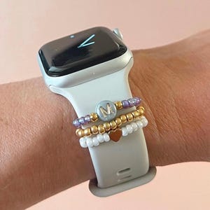 May include: A white Apple Watch with a white band. The watch has a black face with a white clock hand. The watch is being worn on a wrist with three beaded bracelets stacked on top of the watch band. The top bracelet is purple, the middle bracelet is gold, and the bottom bracelet is white. The top bracelet has a silver charm with the letter "M" on it. The middle bracelet has a small gold heart charm.