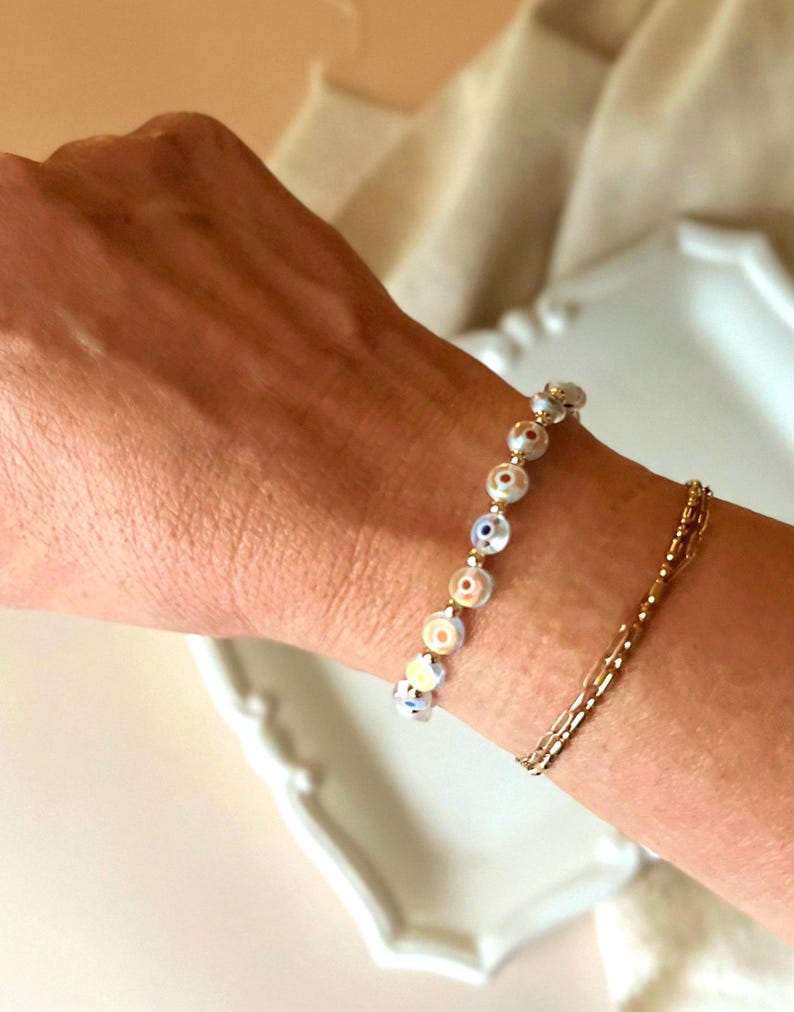 Puede incluir: Un brazalete de cadena dorada con un brazalete de abalorios con peque&ntilde;as cuentas de ojo turco blancas y azules.