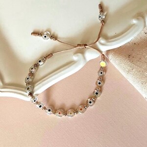 Puede incluir: Un brazalete de cuerda marr&oacute;n con una serie de cuentas de vidrio transparente con un dise&ntilde;o de ojo negro y blanco. Las cuentas est&aacute;n intercaladas con peque&ntilde;as cuentas doradas.