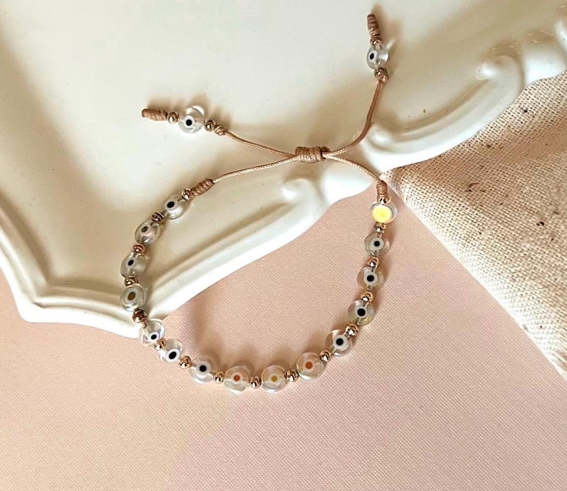 Puede incluir: Un brazalete de cuerda marr&oacute;n con una serie de cuentas de vidrio transparente con un dise&ntilde;o de ojo negro y blanco. Las cuentas est&aacute;n intercaladas con peque&ntilde;as cuentas doradas.