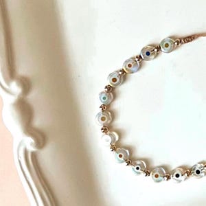 Puede incluir: Un brazalete con una cadena dorada y cuentas blancas con dise&ntilde;os de ojo turco de colores.