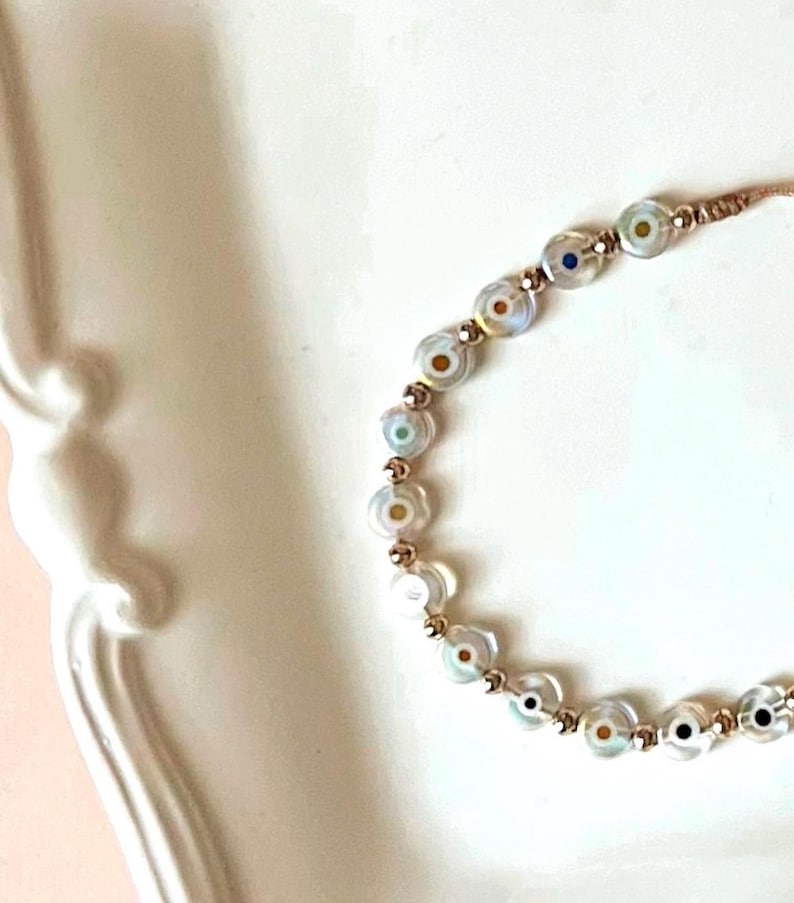 Puede incluir: Un brazalete con una cadena dorada y cuentas blancas con dise&ntilde;os de ojo turco de colores.