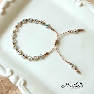 Puede incluir: Un delicado brazalete con una cadena de peque&ntilde;as cuentas azules y blancas iridiscentes en forma de ojo turco. El brazalete es ajustable con un nudo deslizante.