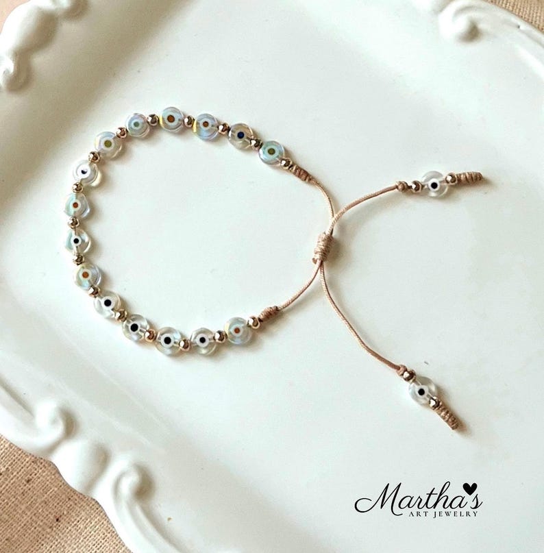 Puede incluir: Un delicado brazalete con una cadena de peque&ntilde;as cuentas azules y blancas iridiscentes en forma de ojo turco. El brazalete es ajustable con un nudo deslizante.