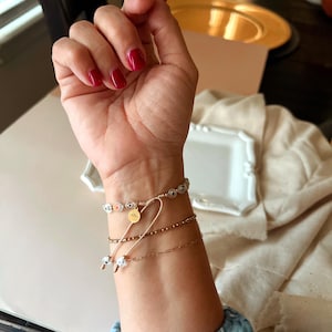 Puede incluir: Una mu&ntilde;eca con tres pulseras. La pulsera superior es una pulsera de cuentas blancas y azules con un dije dorado. La pulsera del medio es una pulsera de cadena dorada. La pulsera inferior es una pulsera de cuentas doradas.