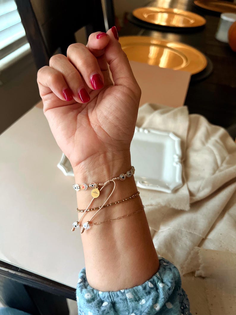 Puede incluir: Una mu&ntilde;eca con tres pulseras. La pulsera superior es una pulsera de cuentas blancas y azules con un dije dorado. La pulsera del medio es una pulsera de cadena dorada. La pulsera inferior es una pulsera de cuentas doradas.