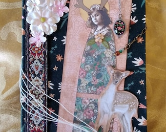 Puede incluir: Un diario hecho a mano con una cubierta de tela floral y estampada. Un marcapáginas decorativo presenta una imagen vintage de una niña con cuernos y un ciervo. Los adornos incluyen flores de papel, cuentas y un clip de metal.